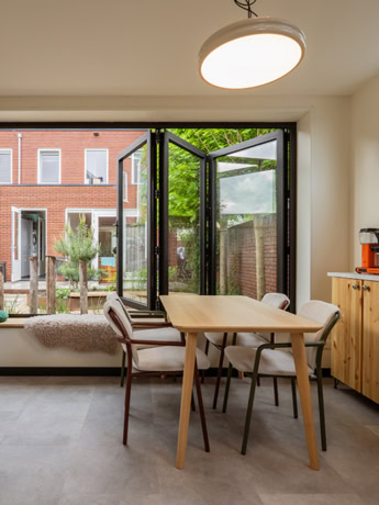 Binnenzicht van een moderne stalen tuindeur met grote ramen, die openstaat naar een tuin met groen gras en bomen, in een lichte en gezellige eetkamer.