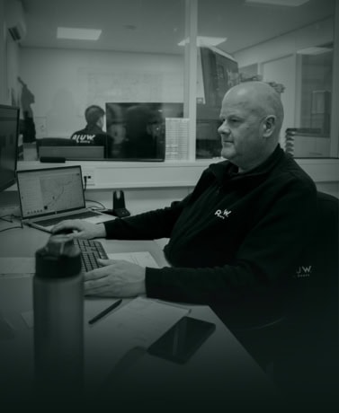 Kantoor medewerker van Aluwdoors die werkt aan projectplannen voor stalen deuren en wanden, met computers en communicatiemiddelen op bureau.