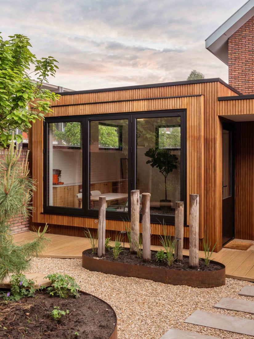 Moderne houten aanbouw met grote glazen ramen en een tuin met planten en houten paaltjes, geschikt voor een stijlvolle woning. Perfect voor een tuin of terras.